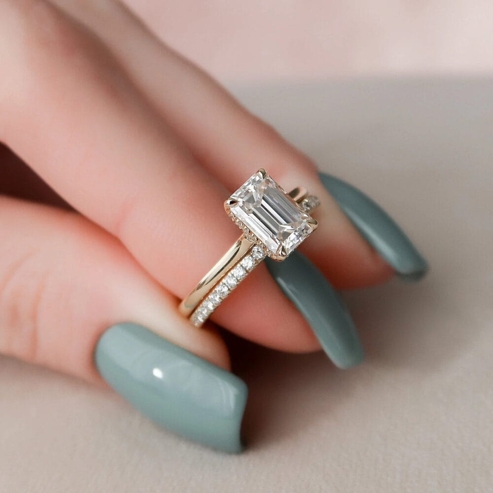 A woman's hand with a teal nail polish wearing a 2.5-carat emerald cut moissanite engagement ring and a matching wedding band. The ring is set in 14k Yellow gold.