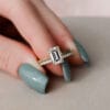 A woman's hand with teal nail polish wearing a 2.5-carat emerald cut moissanite engagement ring and a matching wedding band. The ring is set in 14k yellow gold